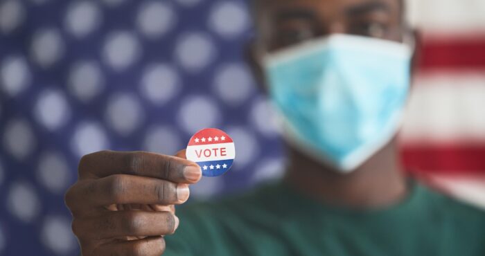 A man wearing a surgial mask standing in front of an American flag holds up a 'vote' sticker.