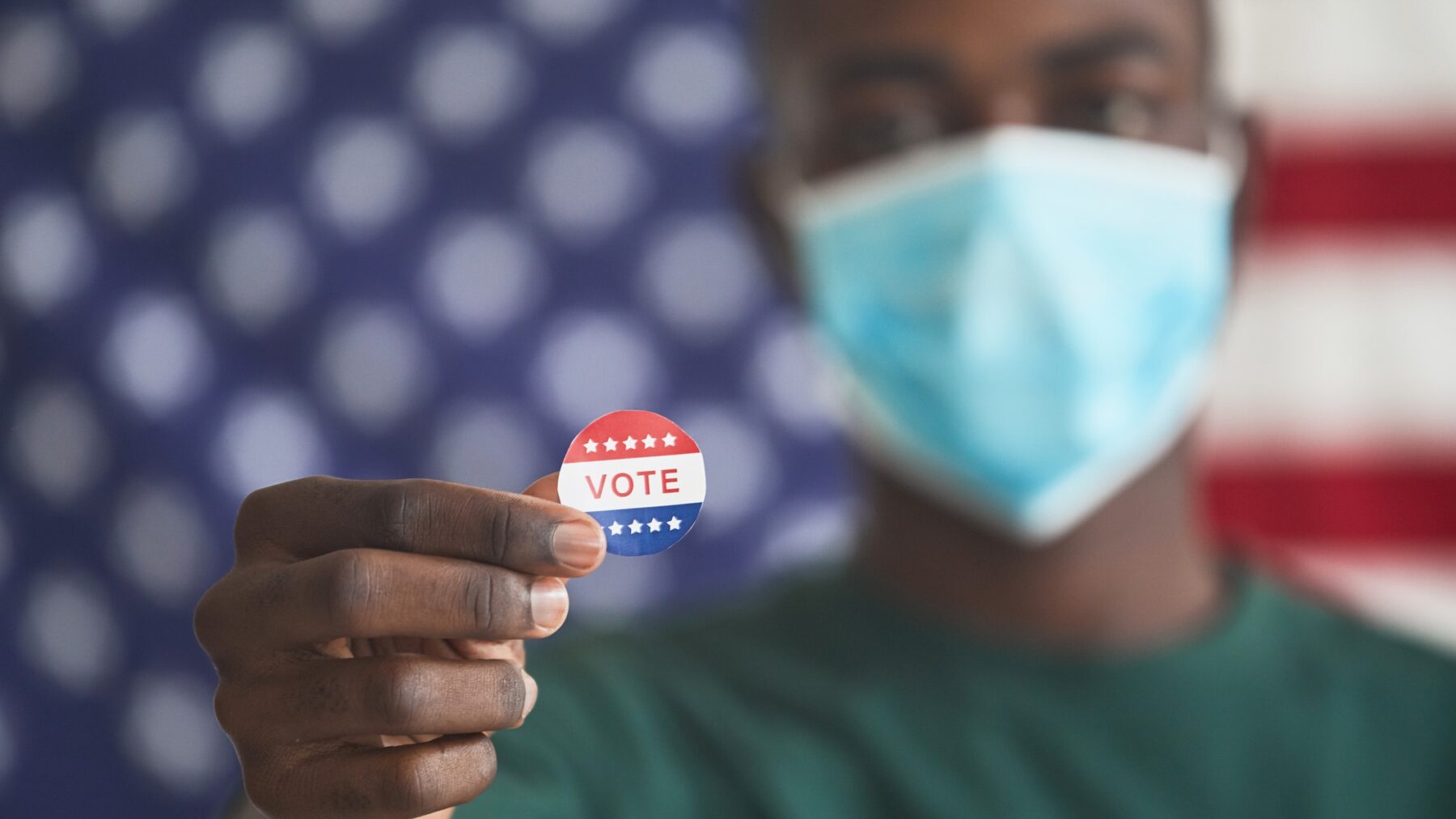 A man wearing a surgial mask standing in front of an American flag holds up a 'vote' sticker.