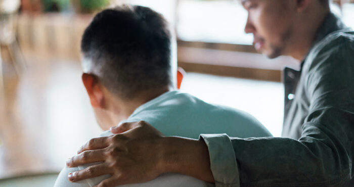 Rear view of son and elderly father sitting together at home. Son caring for his father, putting hand on his shoulder, comforting and consoling him. Family love, bonding, care and confidence