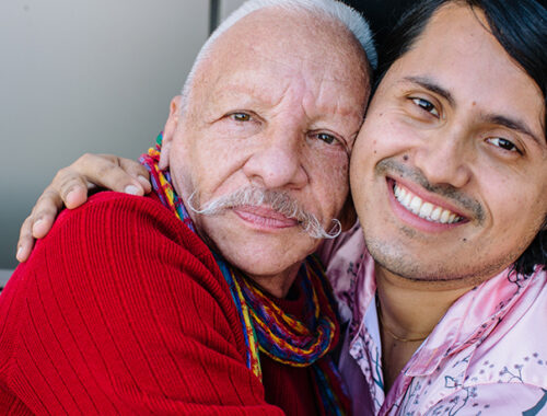 Two Latinx men face the camera, smiling. They have their arms around each other in a hug.