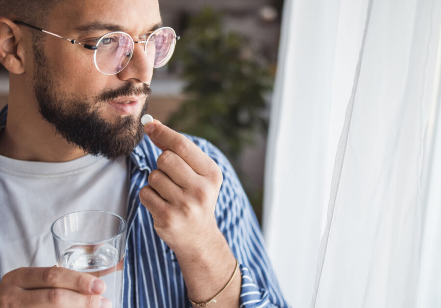 Young man standing by a window, with a glass of water in one hand, holding a pill close to his mouth with the other hand.