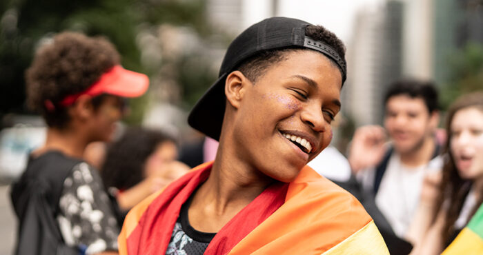Young Black teenager outside surrounded by three other young people in the background; the boy in the foreground looks over his left shoulder, smiling, and is draped in a Pride flag over his shoulders.