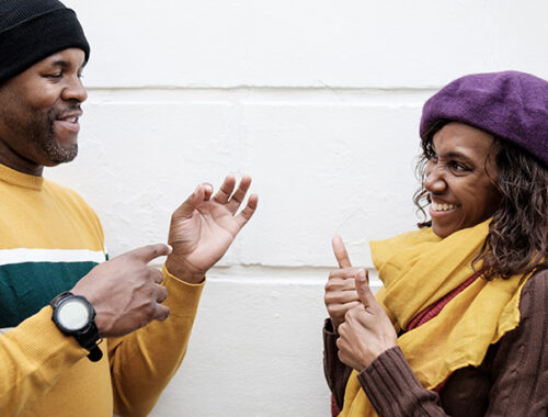 A couple of deaf friends are using sign language in front of a wall background. The wall is white and they are smiling. Autumn weather, they are wearing caps.