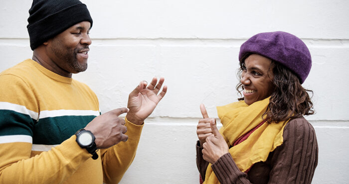 A couple of deaf friends are using sign language in front of a wall background. The wall is white and they are smiling. Autumn weather, they are wearing caps.