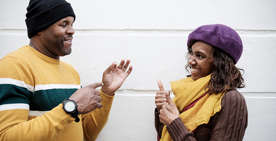 A couple of deaf friends are using sign language in front of a wall background. The wall is white and they are smiling. Autumn weather, they are wearing caps.