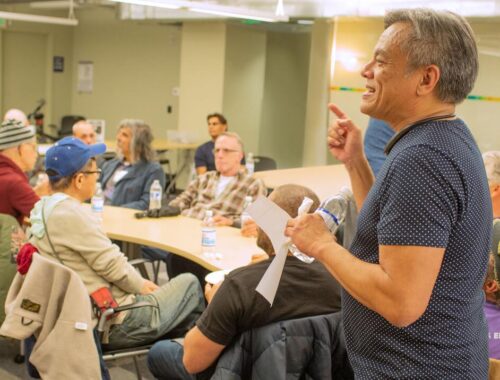 Vince Crisostomo stands at front of room speaking to a group of seniors. He is smiling and looks engaged.