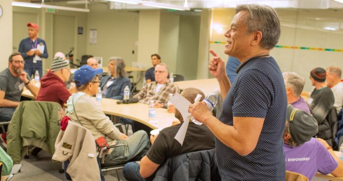 Vince Crisostomo stands at front of room speaking to a group of seniors. He is smiling and looks engaged.