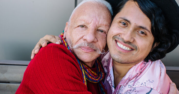 Two Latinx men face the camera, smiling. They have their arms around each other in a hug.