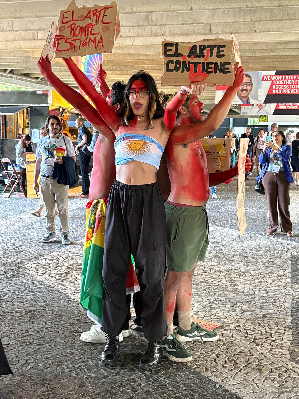 Demonstrators in red body paint hold cardboard signs reading "El arte contiene" during a protest at AIDS 2026