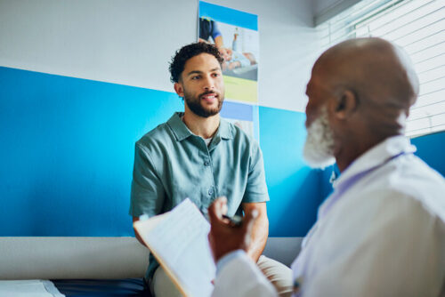 A young Latino man sits in a doctor's office as a doctor sits in front of him, holding a paper chart.