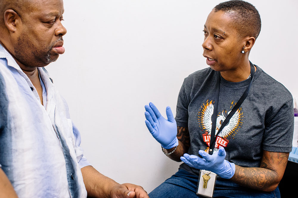 A health worker in gloves and a Live Free From Hep C shirt talks with a client in an exam room