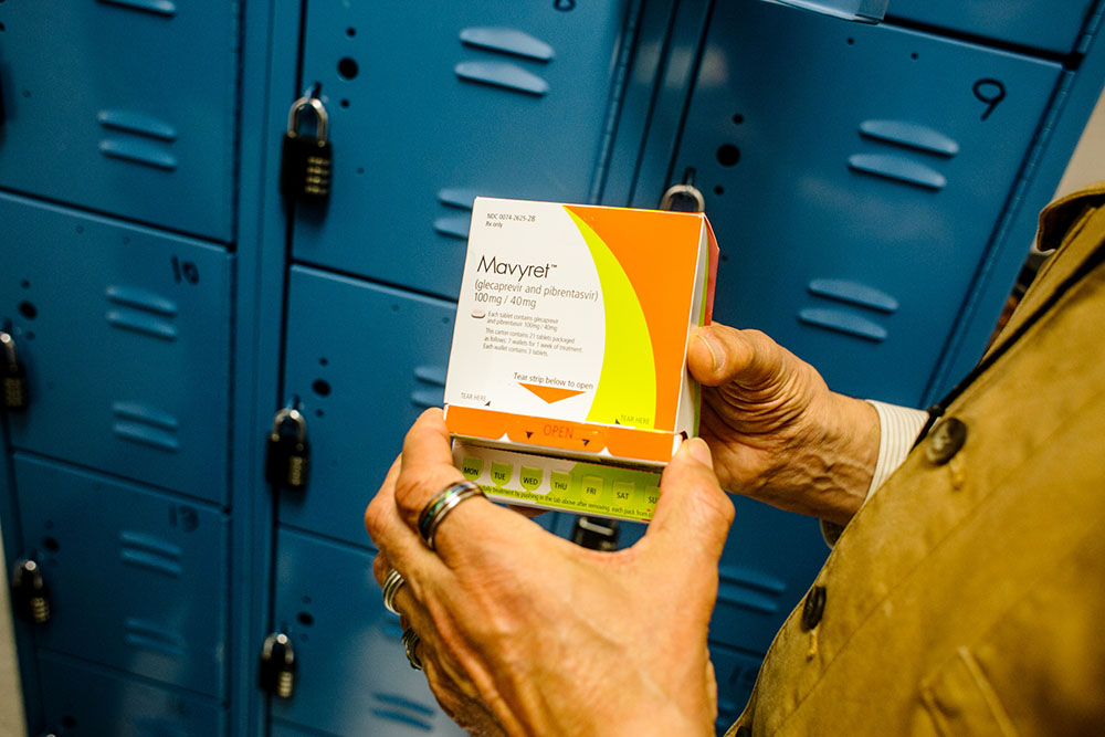 Hands hold a box of Mavyret, a hepatitis C cure medication, in front of blue lockers