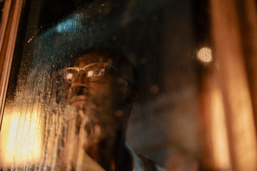 Un hombre con gafas mira por una ventana salpicada por la lluvia por la noche, con la cara iluminada por la cálida luz de las farolas